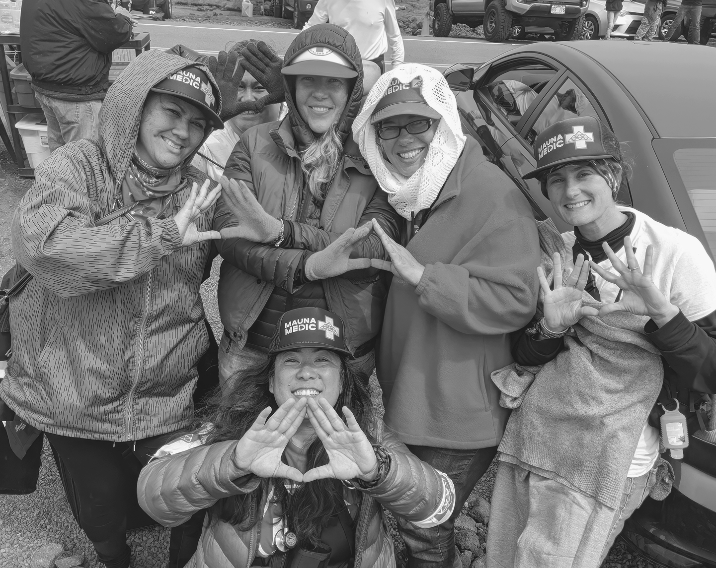 Six women forming interconnected shapes by joining their thumbs and index fingers together with palms facing outward, most wearing Mauna Medic caps, symbolizing unity and solidarity for Maui Medic Healers Hui, a Tides Foundation WE LEAD grantee partner.