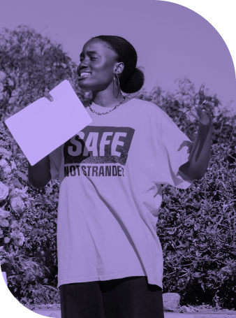 Young Black woman speaks passionately at an outdoor event, holding a sheet of paper and wearing a shirt that reads “SAFE NOT STRANDED,” advocating for safety, dignity, and justice for all communities.