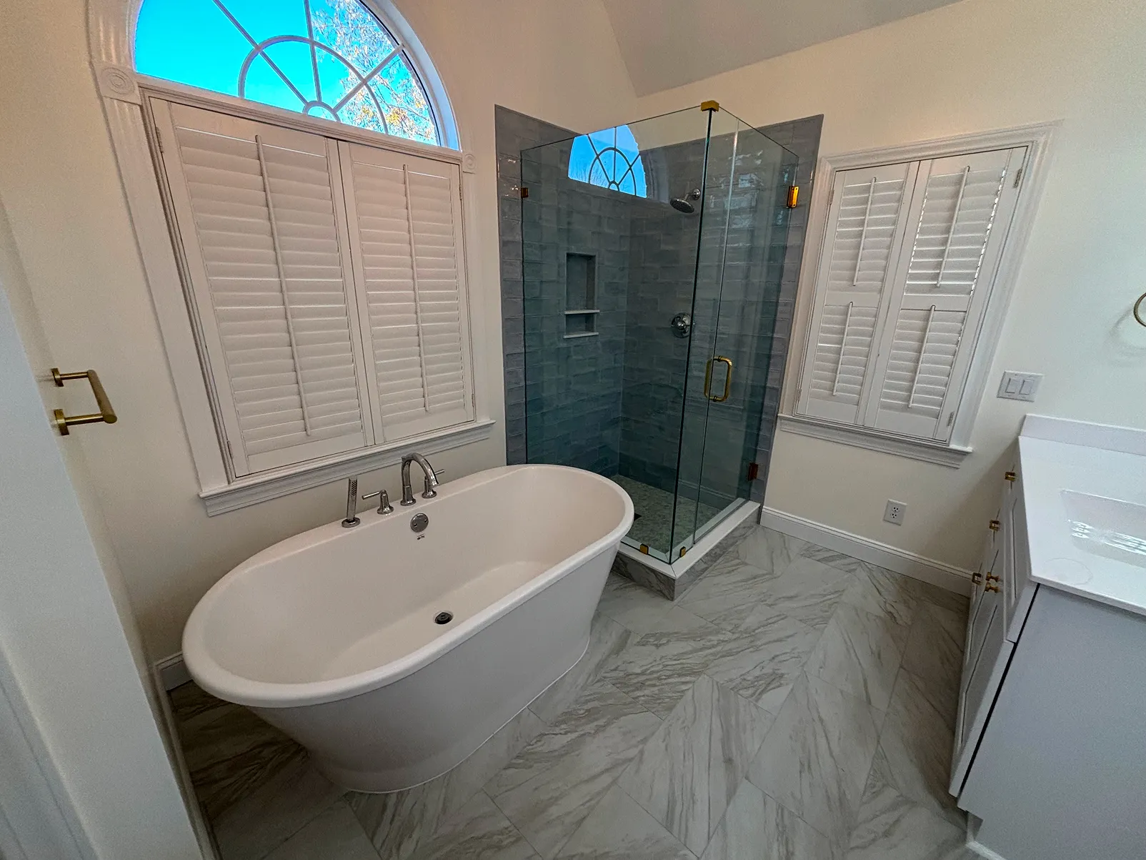 Modern bathroom with a white freestanding bathtub, glass-enclosed shower with blue tile, arched window with shutters, and marble floor tiles.
