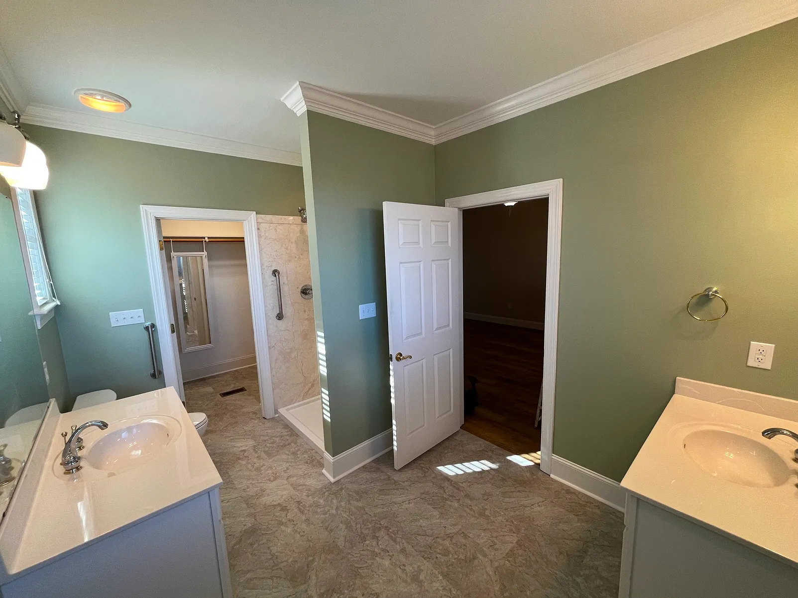 Spacious bathroom with green walls, two white sinks, a walk-in shower, and an open door leading to a room with wooden flooring.