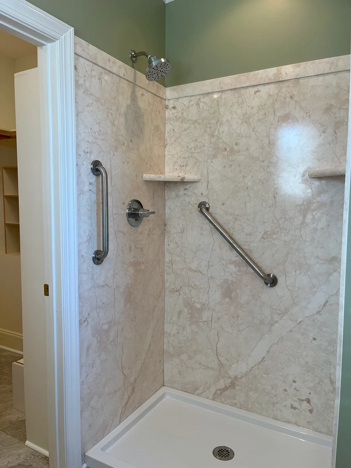 Corner shower with beige marble walls, two metal grab bars, two small corner shelves, a silver showerhead, and a white shower base with drain.