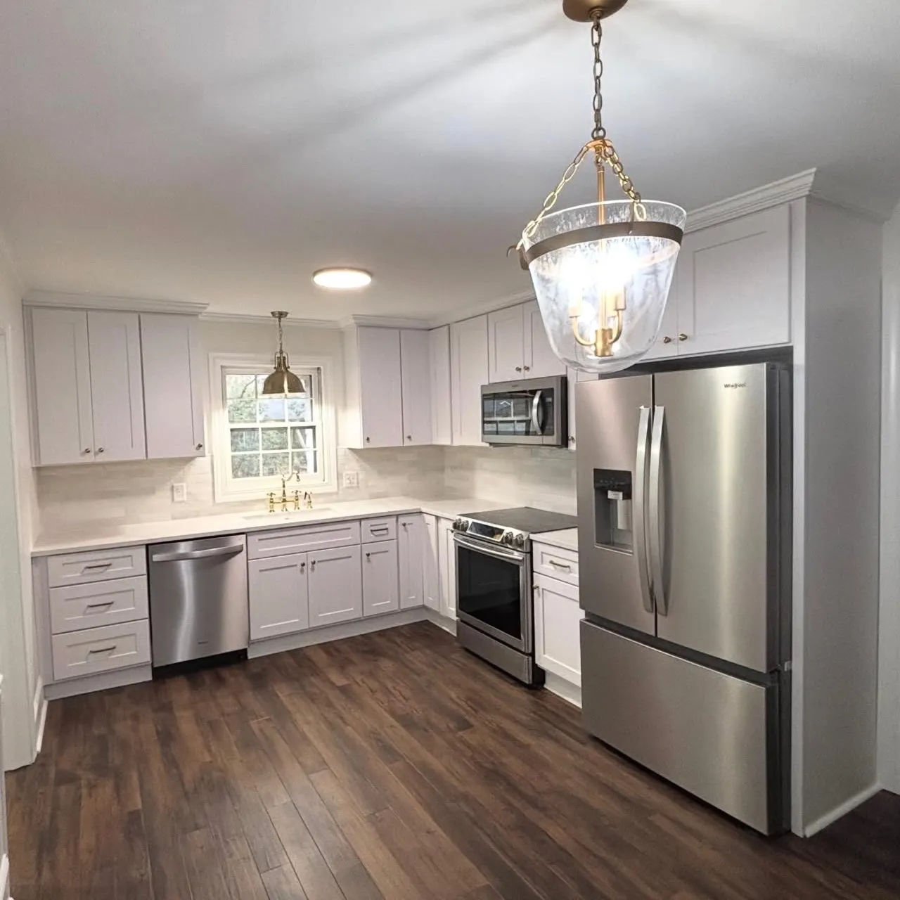 Modern kitchen with white cabinets, stainless steel refrigerator, dishwasher, oven, microwave, brass fixtures, and dark wood flooring.