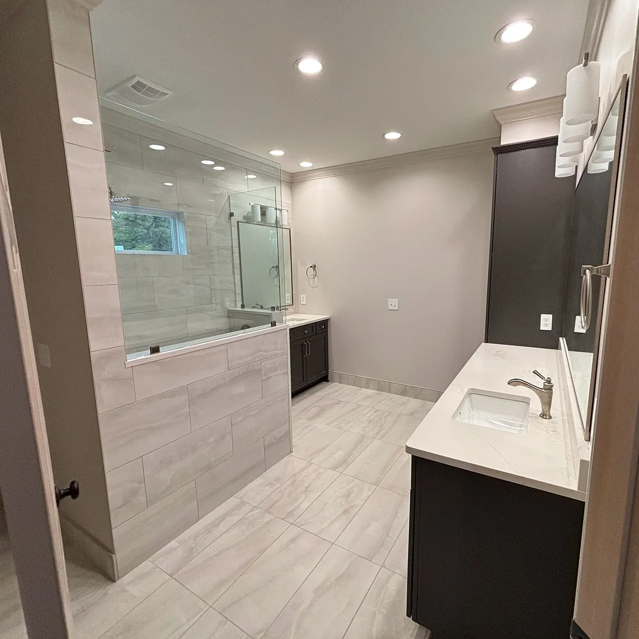Modern bathroom with large beige tile flooring, dual dark wood vanities with white countertops, glass-enclosed walk-in shower, and recessed ceiling lights.