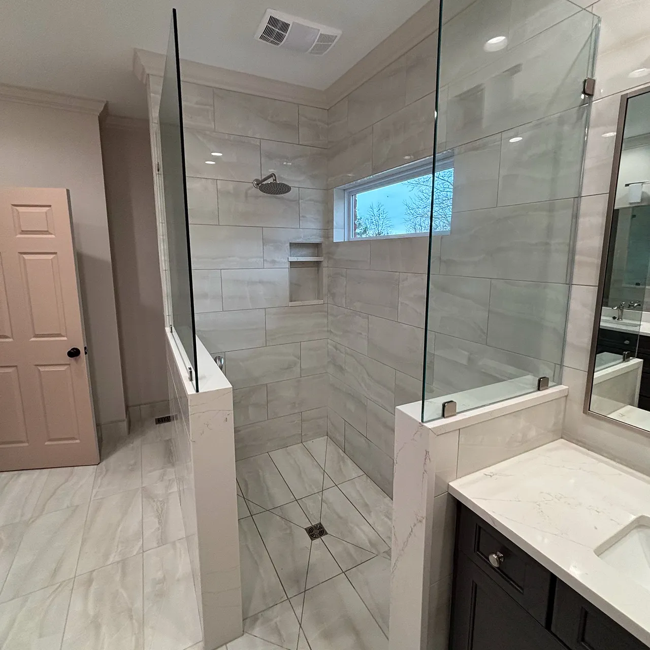 Modern walk-in shower with large gray tiles, glass partial enclosure, a window, and a rain showerhead in a bathroom with light-colored marble floor and vanity.