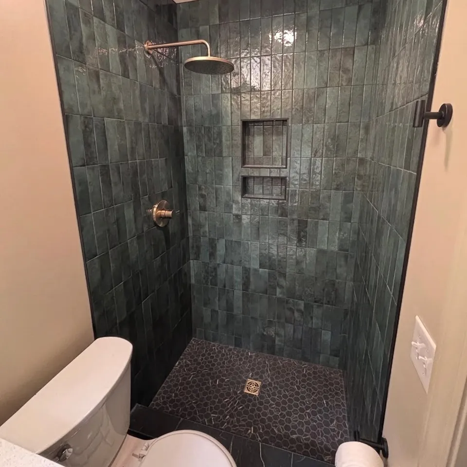 Modern shower area with dark green rectangular wall tiles, small hexagonal floor tiles, wall niches, and a brass showerhead; a toilet and towel holder are visible outside the shower.