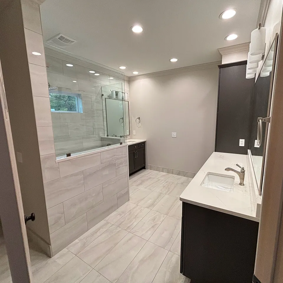 Modern bathroom with light gray tiled floor and walls, dual dark wood vanities with white countertops and recessed ceiling lights.