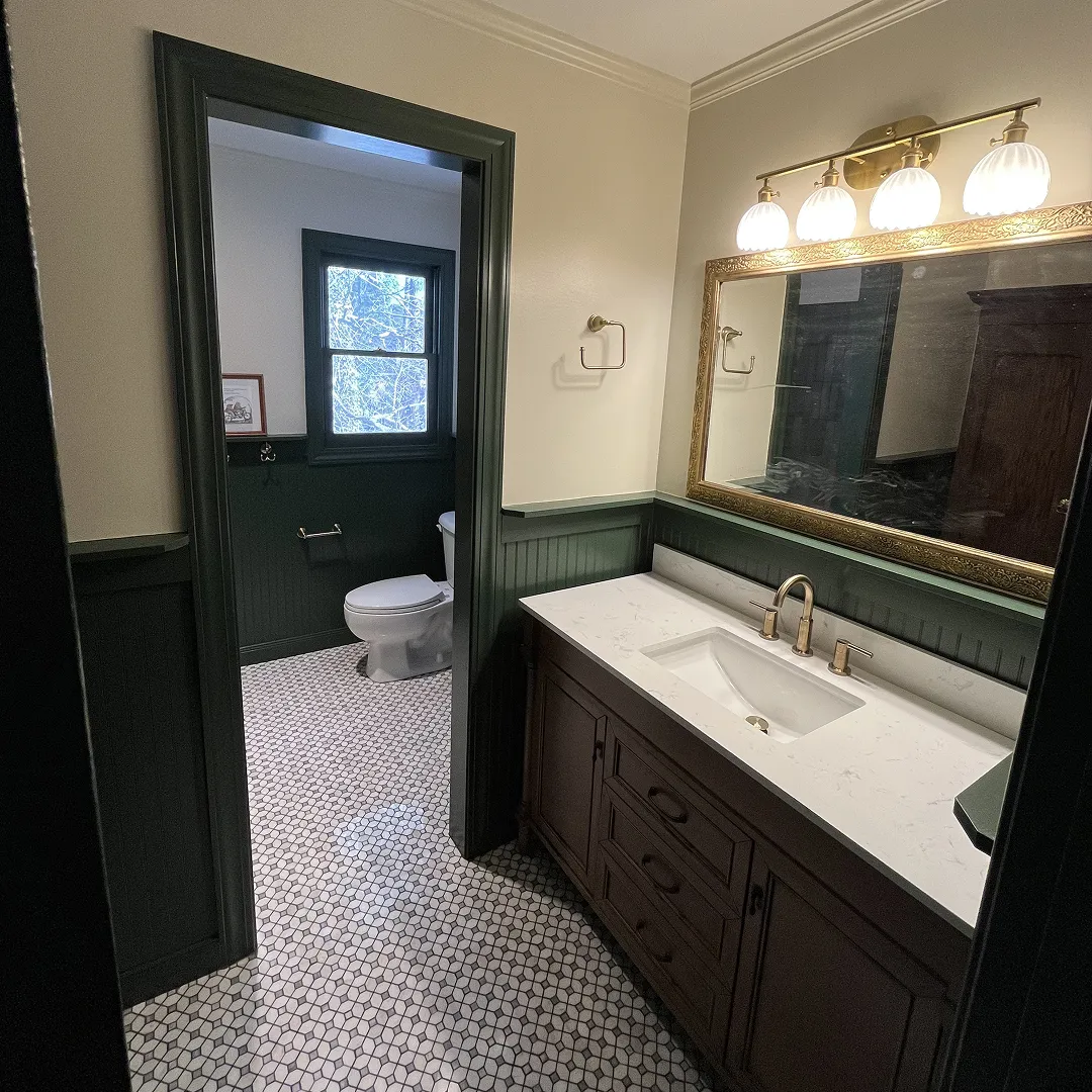 Bathroom with dark green wainscoting, white hexagonal tile floor, wooden vanity with white countertop and brass fixtures, and a toilet next to a window.