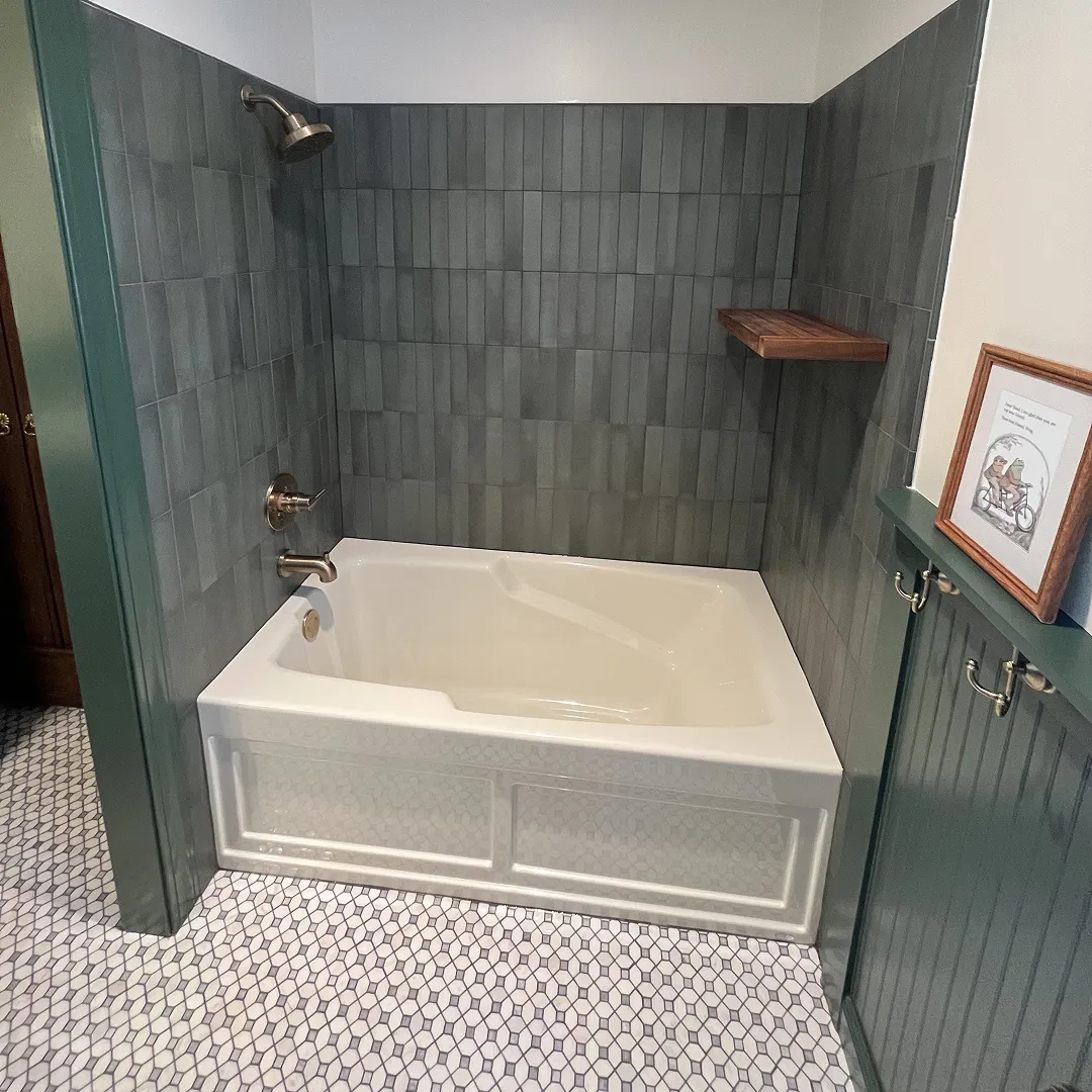 White bathtub with silver fixtures, surrounded by greenish-gray vertical tiles and green wainscoting with hooks, and a framed picture on the right.