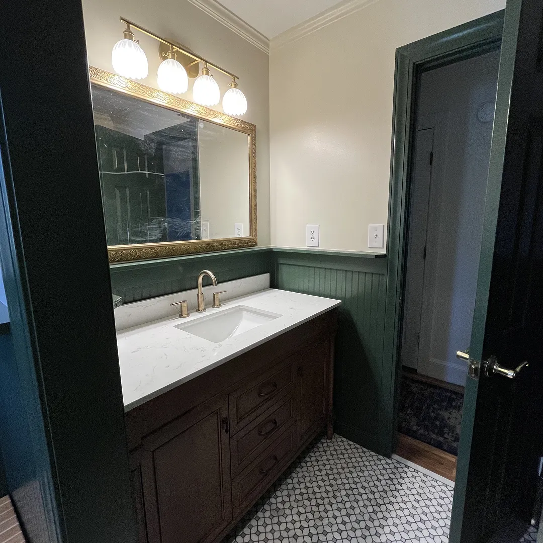 Bathroom sink with white countertop, gold faucet, wooden cabinet, large gold-framed mirror, and green wainscoting.