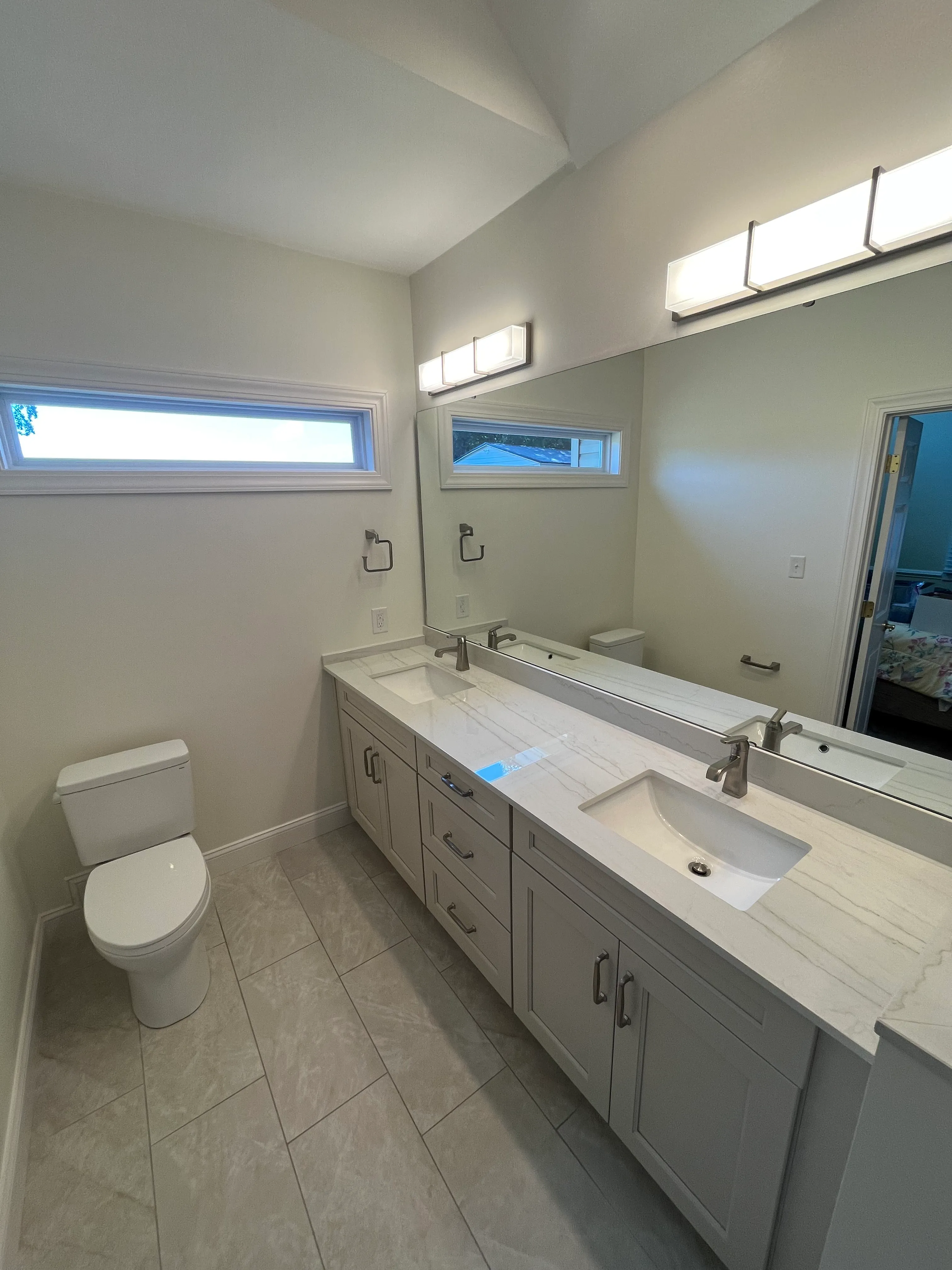 Modern bathroom with dual sinks, large mirror, white cabinets, toilet, and gray tiled floor.