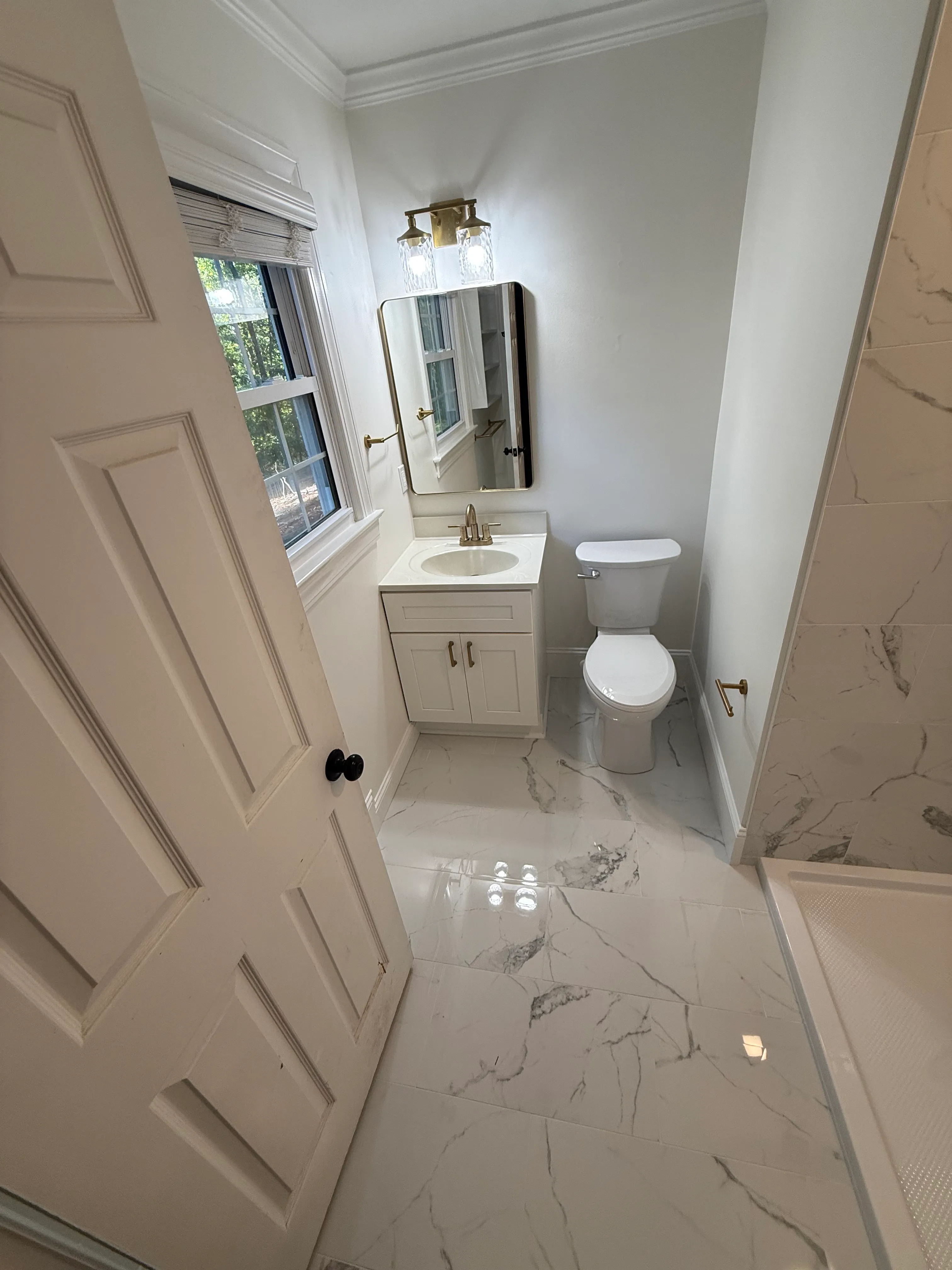 Small bathroom with white marble tiled floor, white wooden door partially open, window with white blinds, white vanity with sink and brass faucet, rectangular mirror with brass frame, two-bulb brass light fixture above, white toilet, and a corner shower with marble tiled walls.