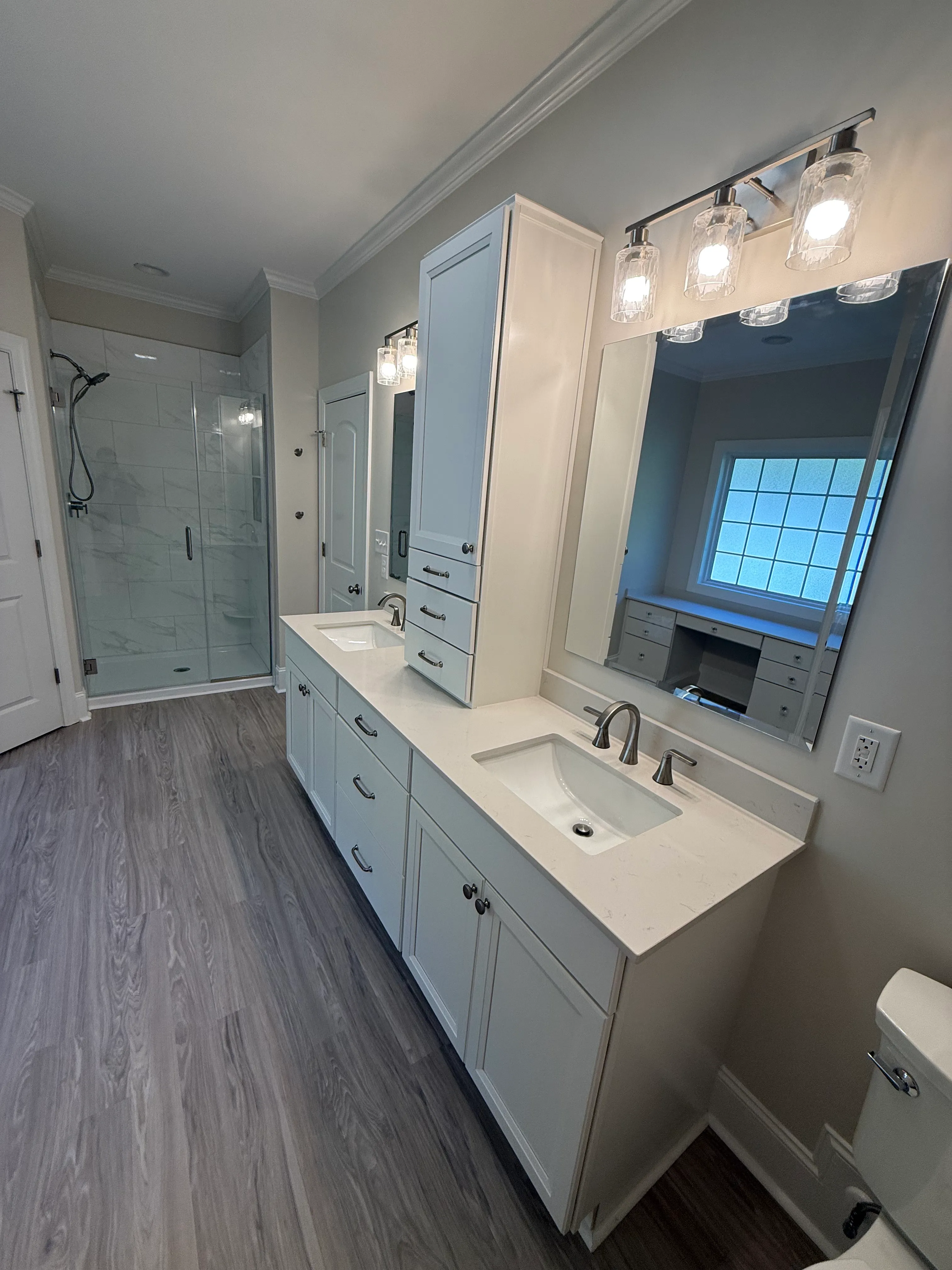 Modern bathroom featuring a double sink vanity with white cabinets and quartz countertop, large rectangular mirror with three polished metal and glass light fixtures above, frosted window reflected in the mirror, wooden plank flooring, glass-enclosed shower with white marble tile and matte black fixtures, and a white toilet partially visible in the foreground.