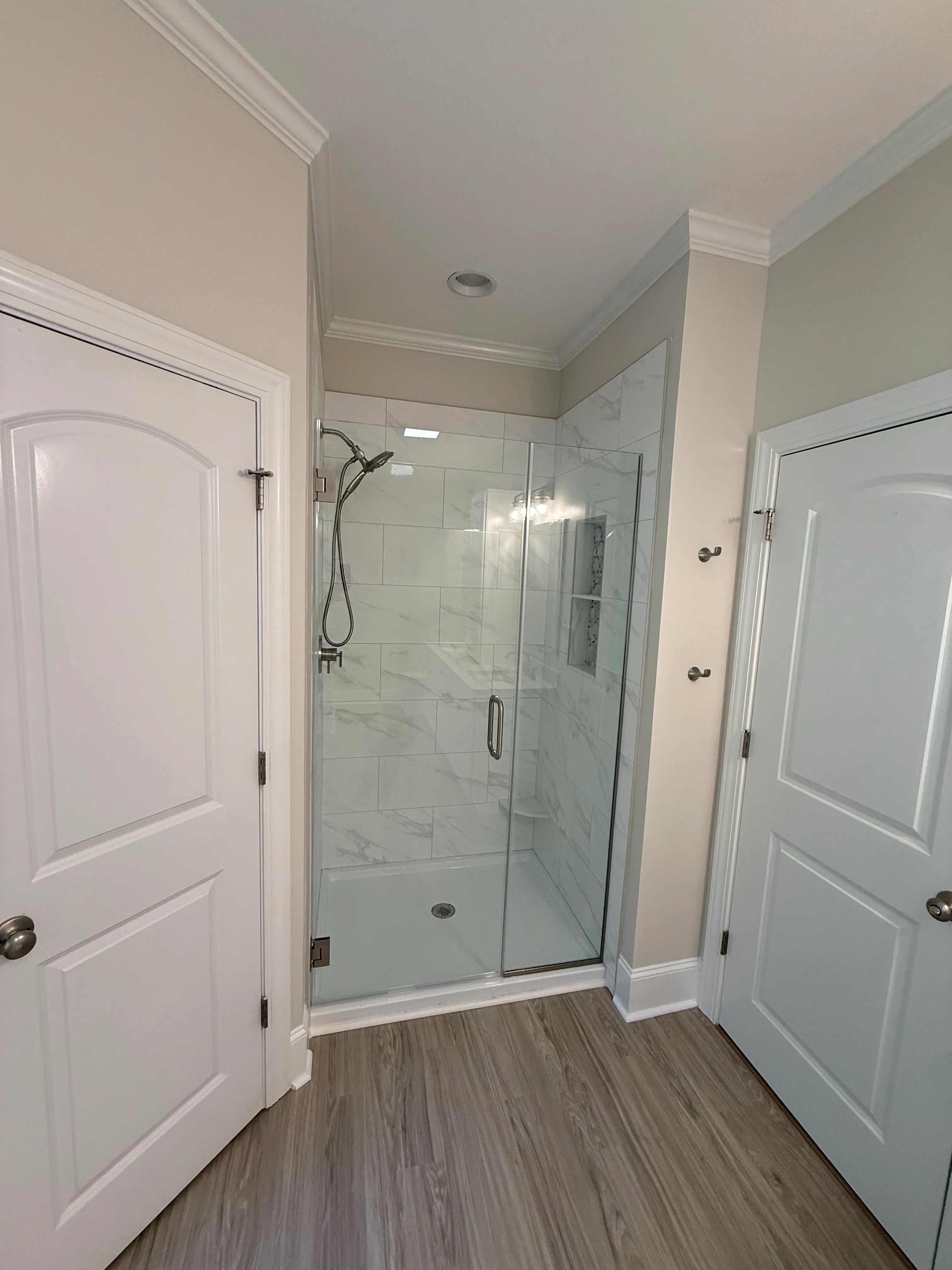 Modern corner shower with glass sliding doors, white and gray marble-style tile walls, a handheld showerhead, and a built-in wall niche, flanked by two white doors and light wood flooring.