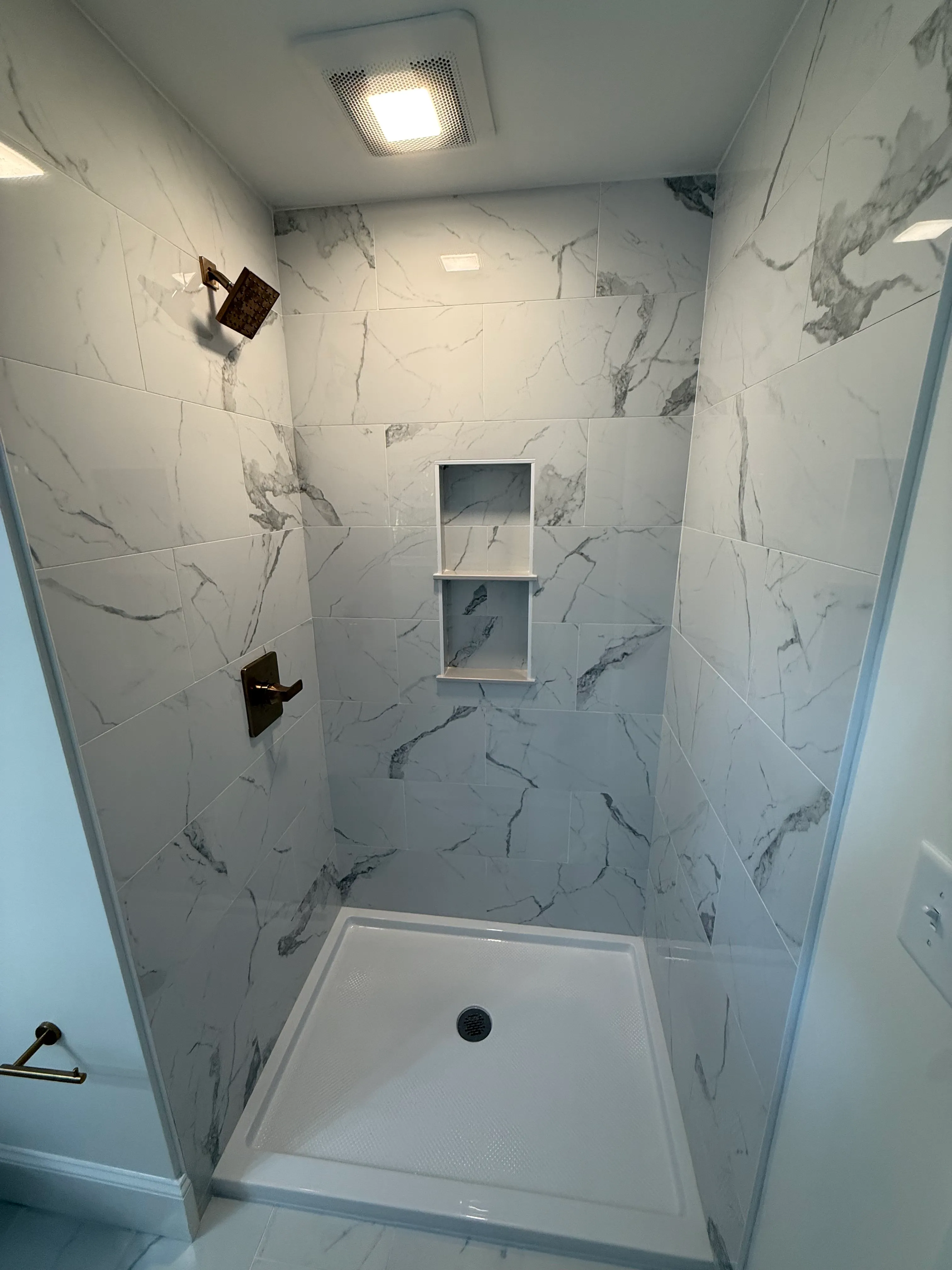 Small shower stall with white marble-effect tiled walls, built-in two-tier shelf, bronze showerhead, bronze faucet handle, and a white textured shower base with a drain in the center.