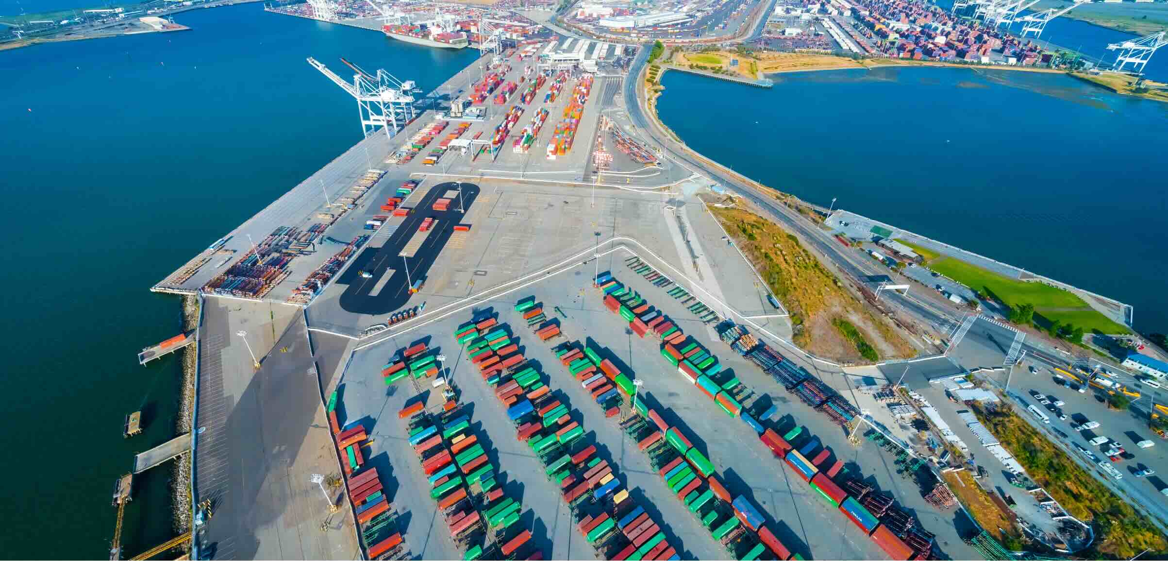 An aerial view of a busy shipping yard featuring rows of colorful shipping containers and cranes near a calm waterway, with urban development visible in the background