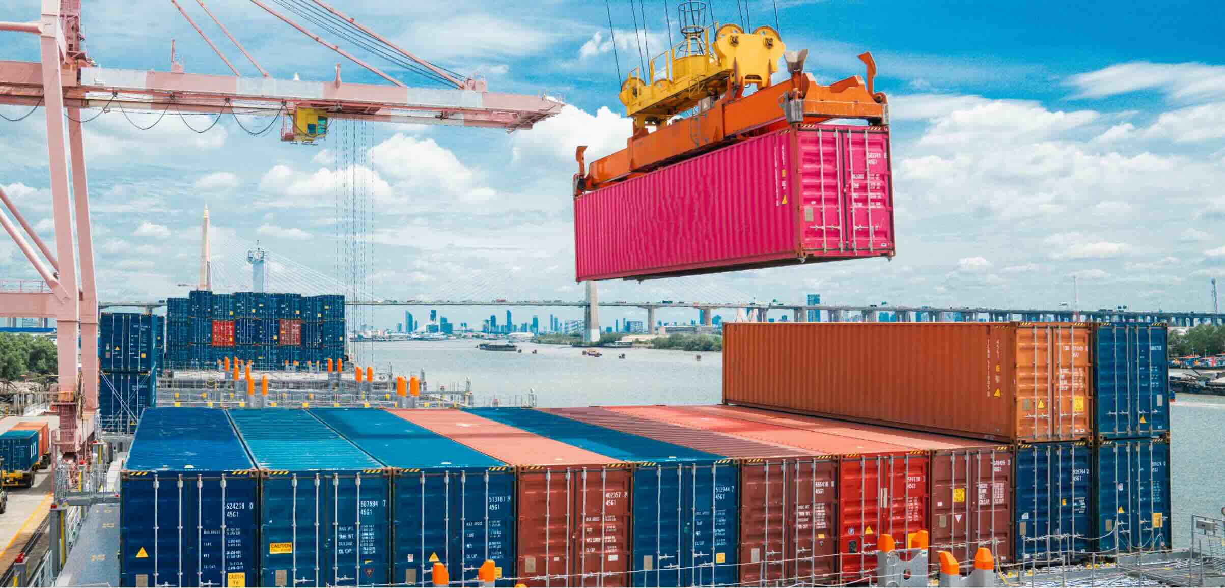 A large pink shipping container being lifted by a crane above stacked cargo containers at a bustling port