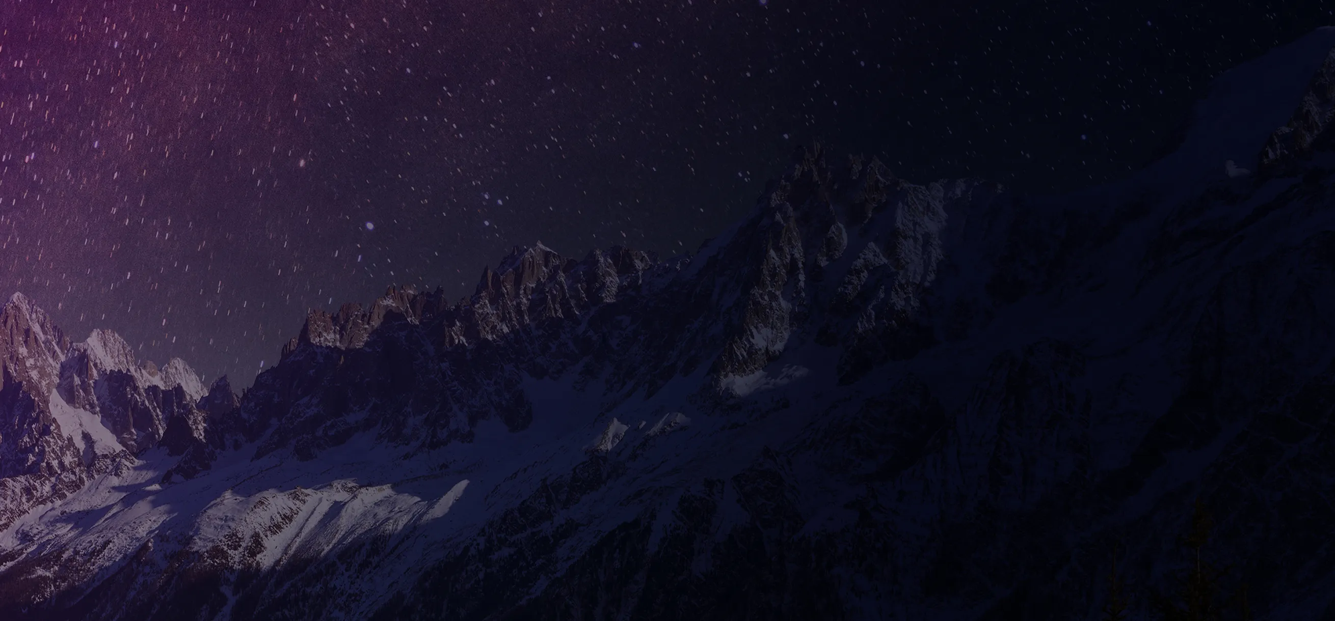 Snow-covered mountain range under a starry night sky with a purplish hue.