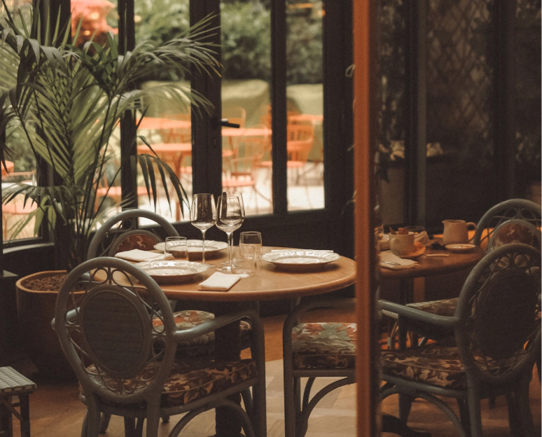 Salle de restaurant de l'hôtel La Fantaisie à Paris 9 avec tables d'hôtes dressées, vaisselle en porcelaine, verres à pied et chaises en rotin tissé devant une verrière lumineuse donnant sur une cour-jardin.