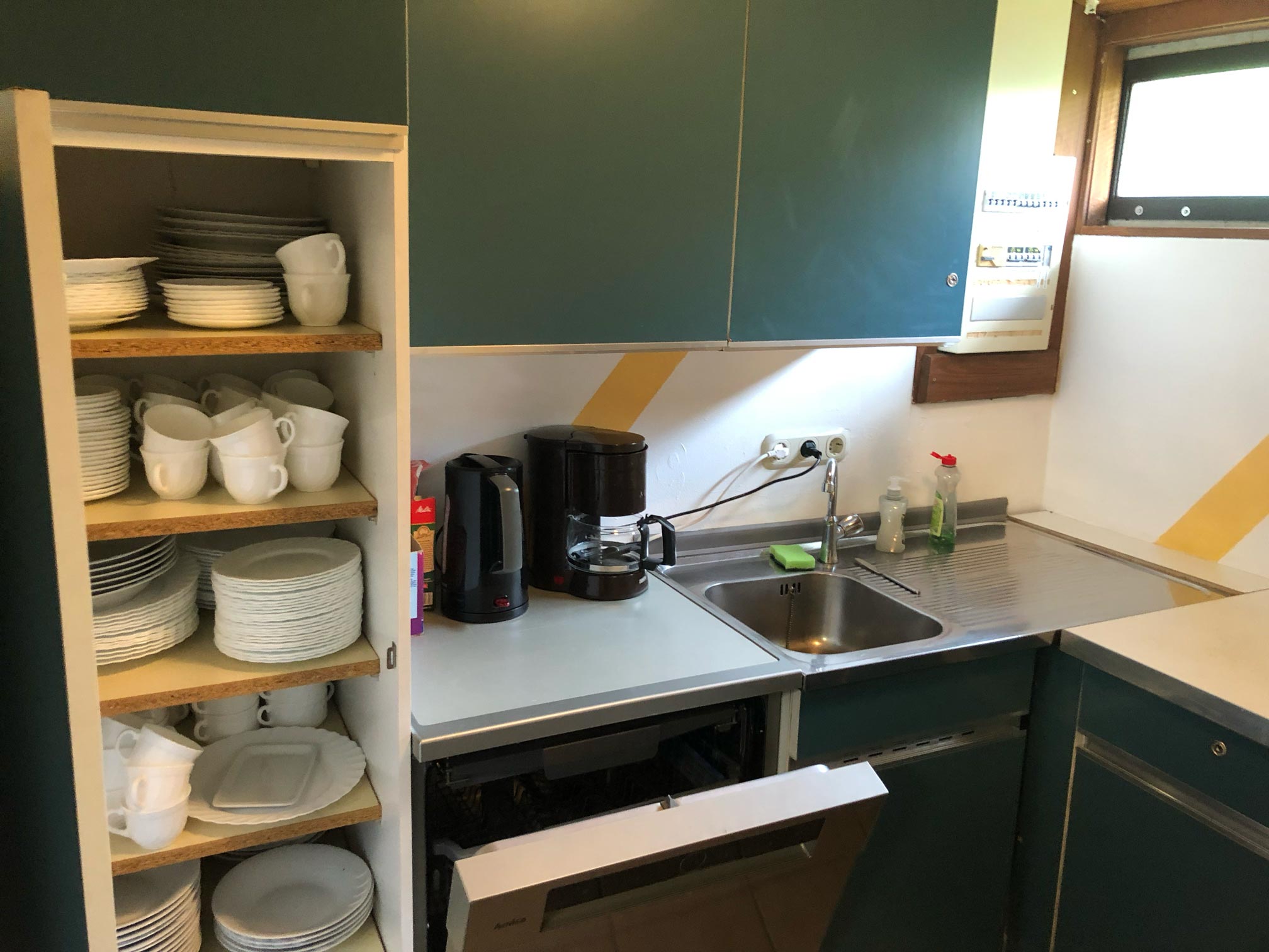 Ansicht der Küche mit türkisfarbenen Fronten, ein großer Schrank ist geöffnet und zeigt innen Geschirr für ca. 40 Personen, eine Spüle, ein Wasserkocher und eine Kaffemaschine sind zu sehen