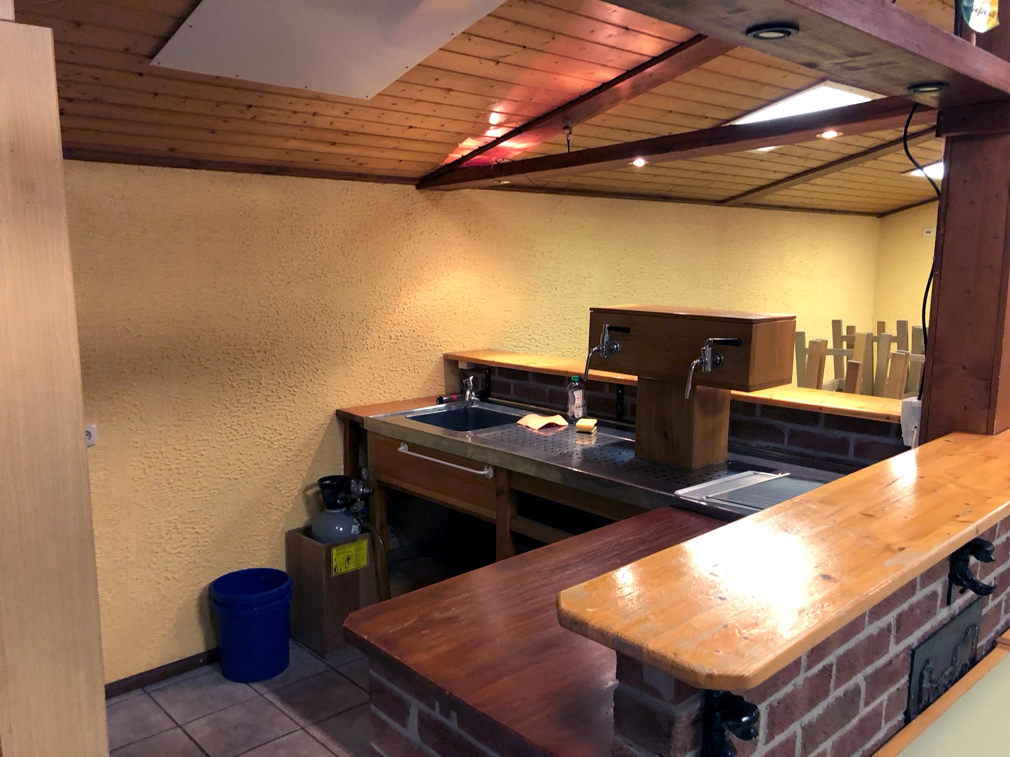 Vereinshaus von innen mit Sicht auf eine gemauerte Theke, Das Dach mit brauner Holzvertäfelung und gelb gestrichenen Wänden