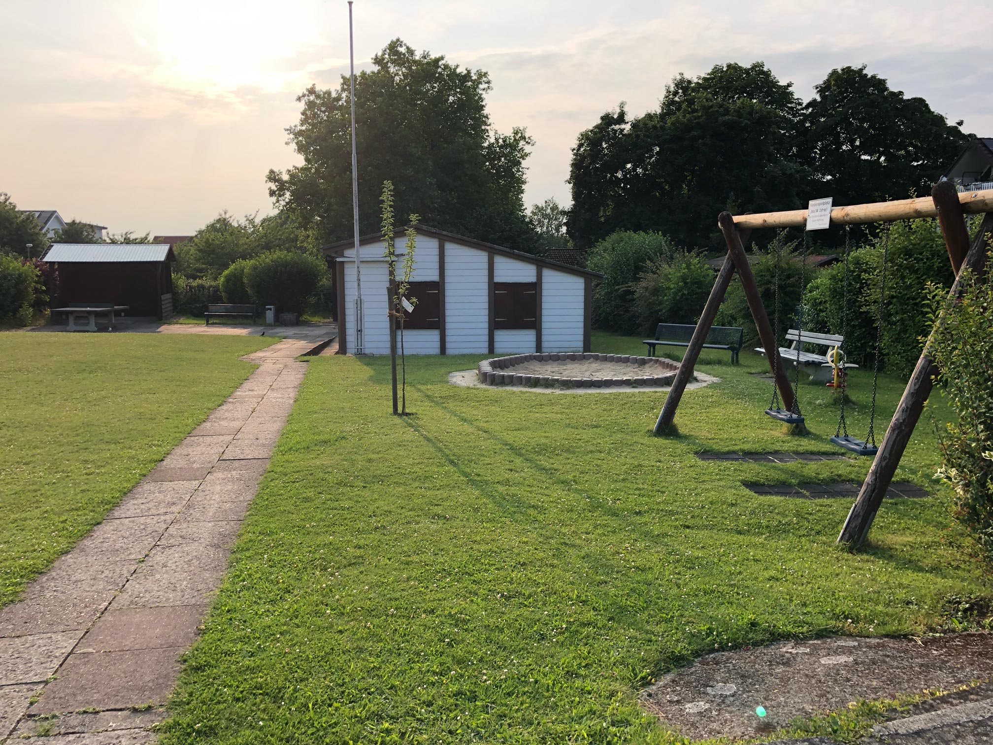 Blick auf eine große Wiese, vorne eine Schauke mit Sandkasten, hinten das Vereinshaus und hinten links eine kleine Überdachung und eine Tischtennisplatte