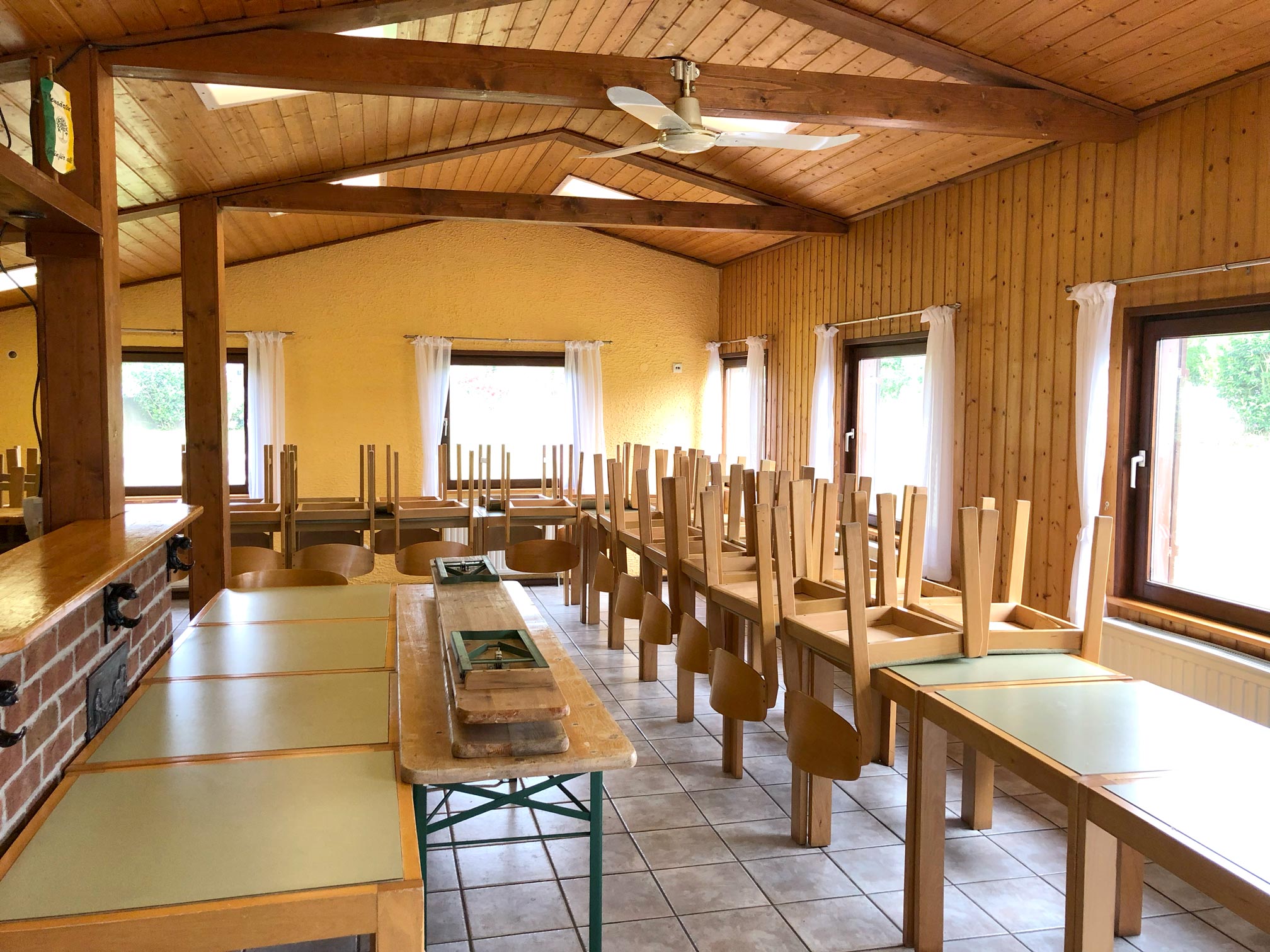 Vereinshaus von innen, Lichtdurchflutete Fenster, das Dach mit brauner Holzvertäfelung und gelb gestrichenen Wänden, Tische stehen im Raum mit aufgestellten Stühlen