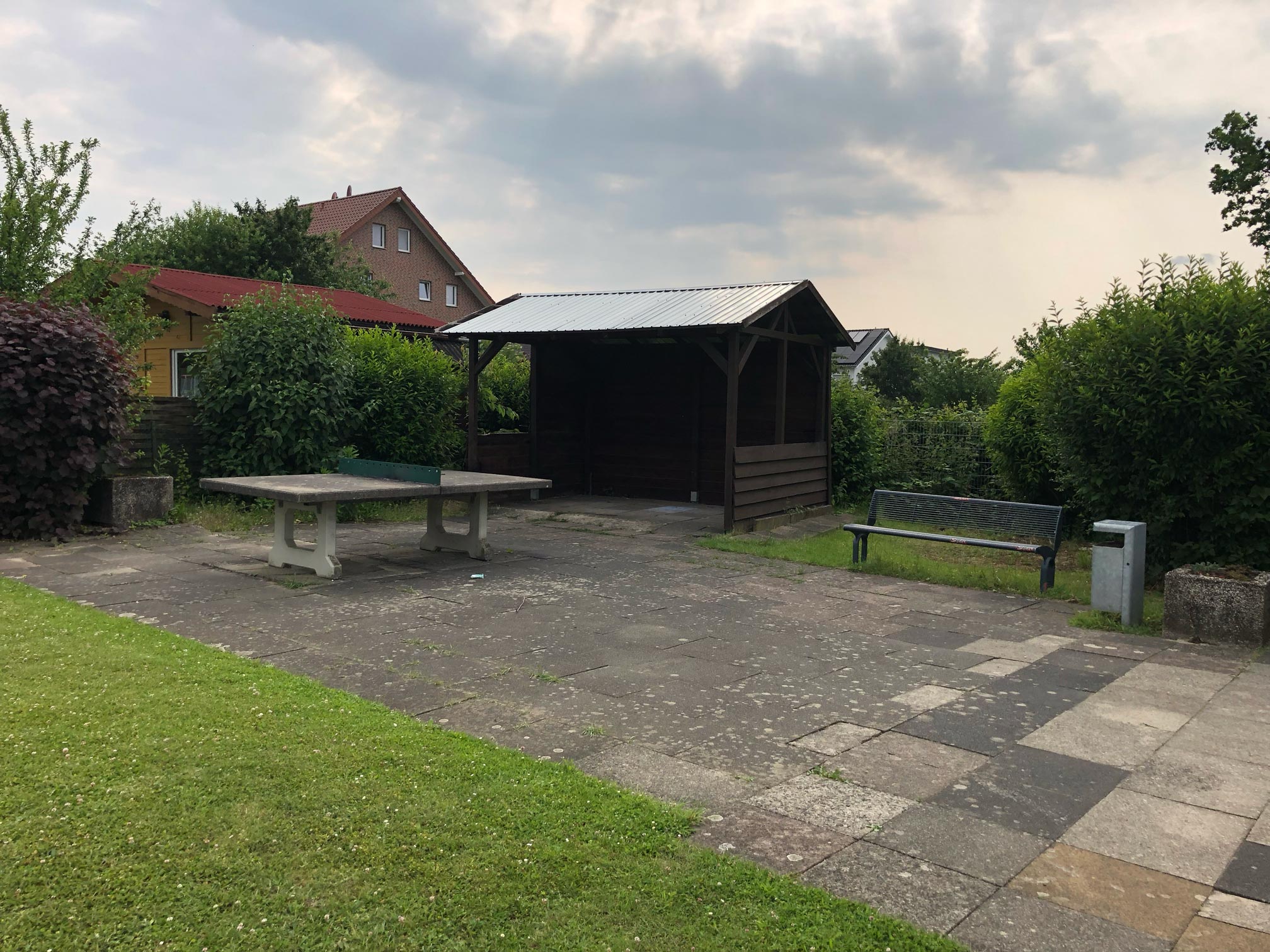 Blick auf eine gepflasterte Fläche, darauf steht eine Tischtennisplatte aus Stein, dahinter eine kleine Überdachung aus Holz