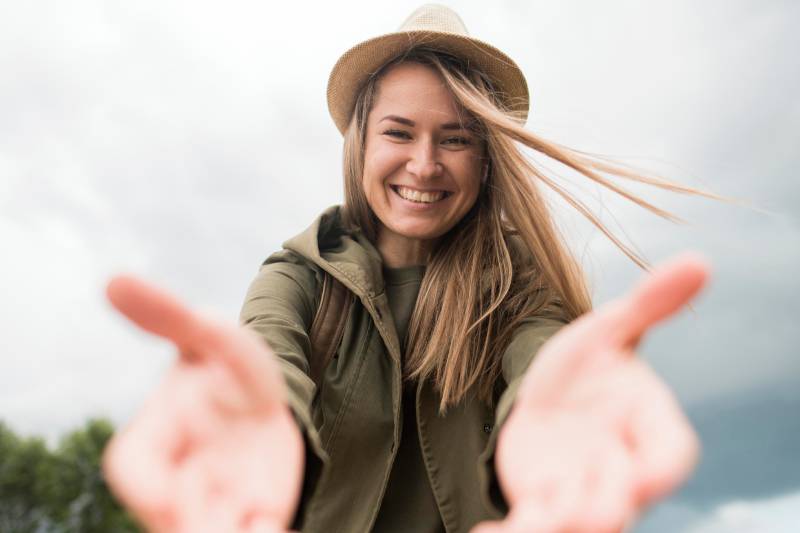 Jeune femme souriante tendant les bras vers l’avant, portant un chapeau et une veste kaki en extérieur.