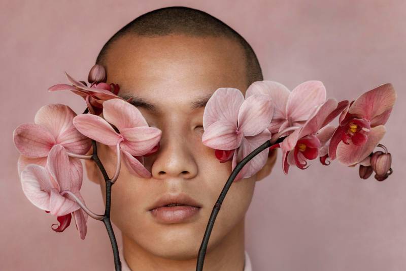 Portrait d’une personne aux yeux fermés, encadrée par des orchidées roses sur fond rose poudré.
