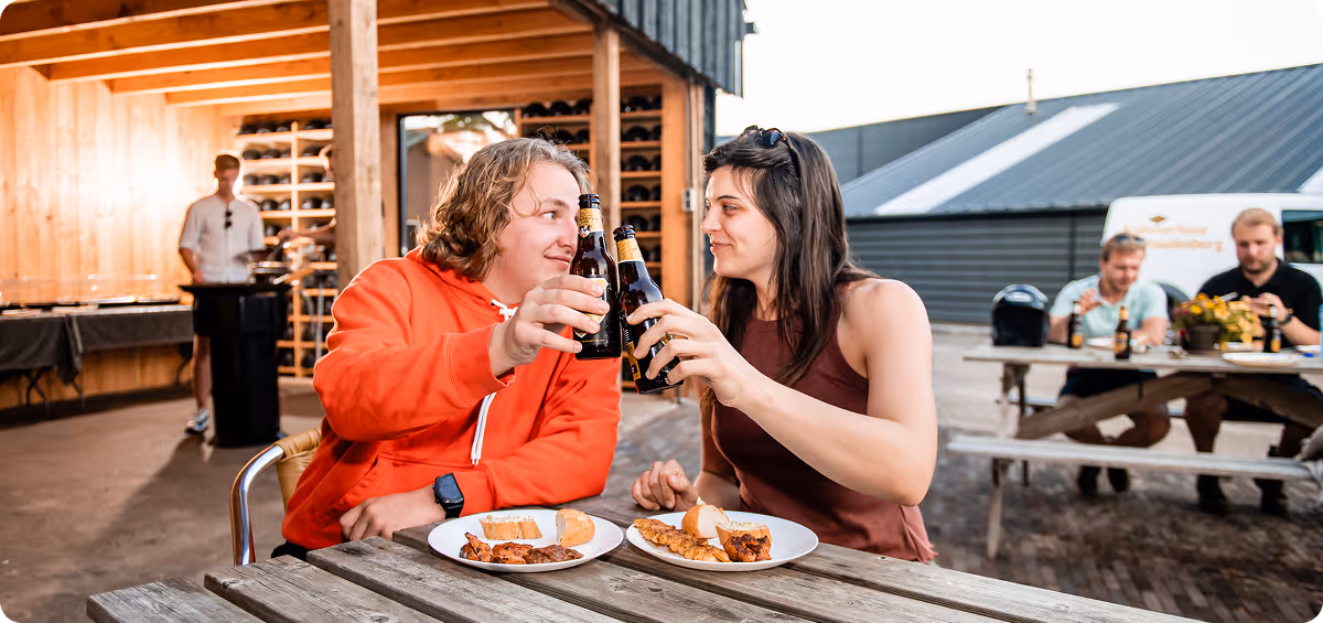 Twee mensen zitten buiten aan een picknicktafel, proosten met bierflesjes en lachen naar elkaar, terwijl ze genieten van een maaltijd met vlees en brood; op de achtergrond zitten andere mensen ook aan tafels.