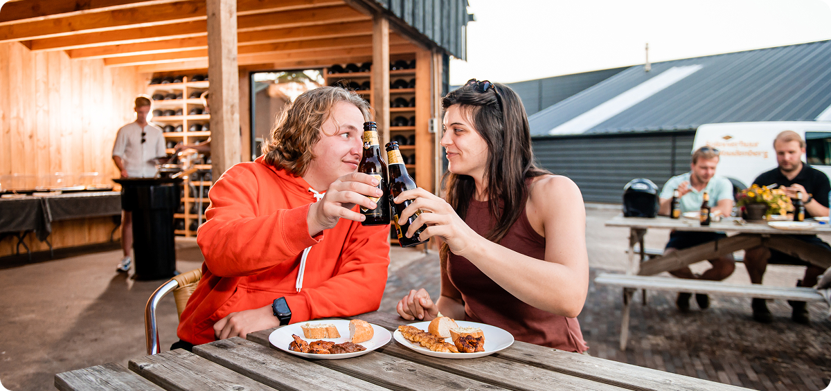 Twee mensen zitten buiten aan een picknicktafel, proosten met bierflesjes en lachen naar elkaar, terwijl ze genieten van een maaltijd met vlees en brood; op de achtergrond zitten andere mensen ook aan tafels.