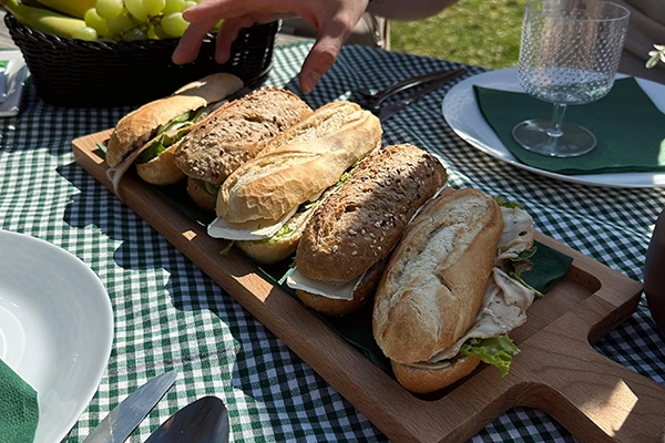 Houten serveerplank met zes belegde broodjes, waaronder volkoren en witte varianten met sla en kipfilet, op een groen-wit geruit tafelkleed tijdens een zonnige buitentafelsetting.