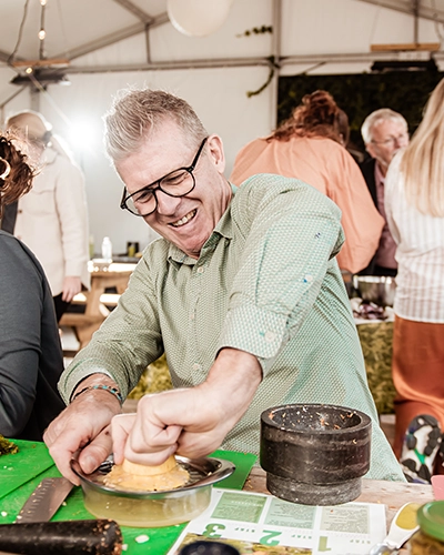 Man met bril en groene blouse perst met enthousiasme een sinaasappel tijdens een workshop in een drukbezochte tent met andere mensen op de achtergrond.