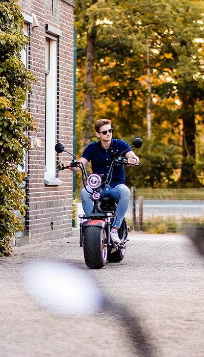 Man met zonnebril rijdt op een brede elektrische chopper langs een bakstenen gebouw, met bomen en een weg op de achtergrond in de avondzon.