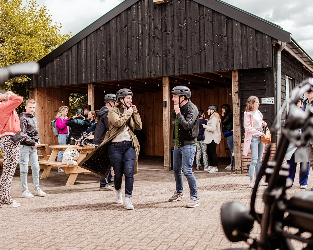 Groep mensen verzamelt zich bij een houten gebouw, waar ze helmen opzetten ter voorbereiding op een activiteit; op de voorgrond staan elektrische steps, en er heerst een levendige en vrolijke sfeer.