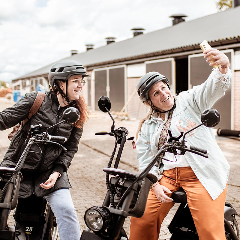 Twee vrouwen met helmen zitten op elektrische steps en lachen terwijl ze samen een selfie maken op een boerenerf met schuren op de achtergrond.