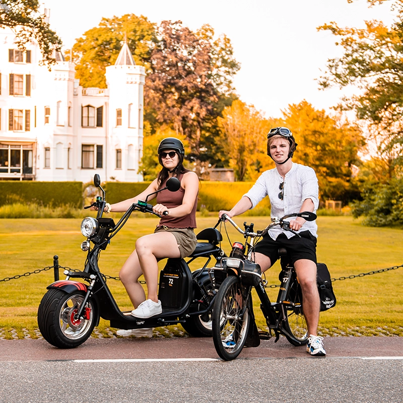 Man en vrouw poseren op hun elektrische steps op een fietspad voor een wit landhuis, omgeven door een groene tuin en bomen in de avondzon.