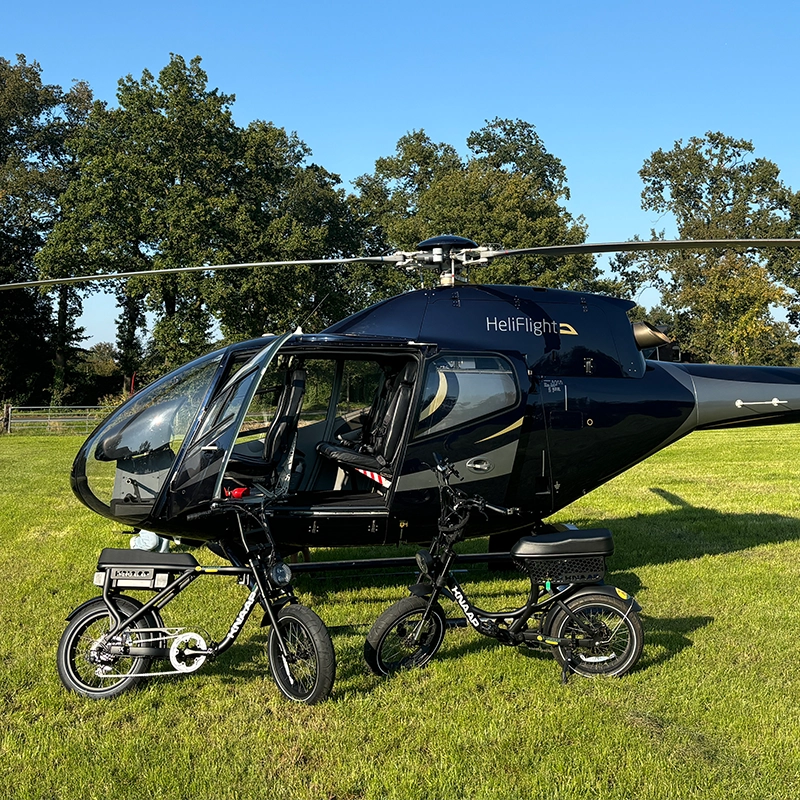 Zwarte helikopter met open deur staat op een grasveld, met twee elektrische fietsen ervoor en bomen op de achtergrond onder een heldere blauwe lucht.