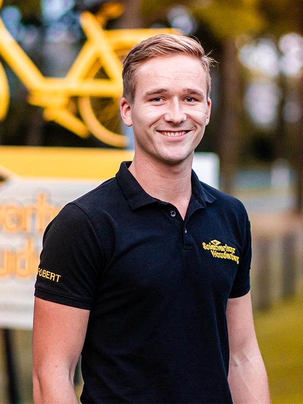 Jonge man met kort blond haar en zwart poloshirt met gele opdruk lacht naar de camera; op de achtergrond staat een geel bord met een fietssymbool en bedrijfsnaam.