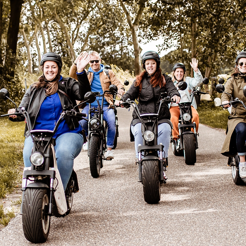 Vrolijke groep mensen rijdt op brede elektrische steps over een boomrijk fietspad; sommigen zwaaien naar de camera terwijl ze genieten van een gezamenlijke rit.