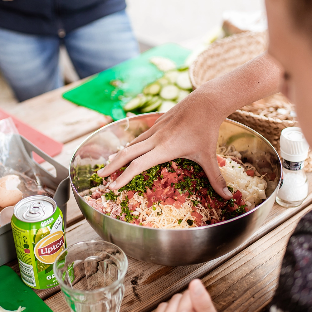 Hand mengt gehakt, ui, tomaat, kaas en verse kruiden in een grote metalen kom op een houten tafel, met kookspullen en een blikje Lipton Ice Tea in beeld.