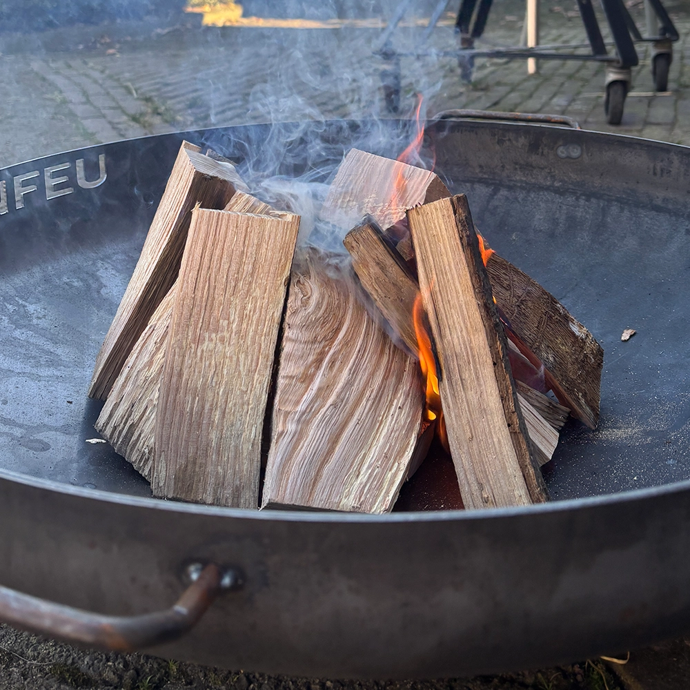 Vuurkorf met stapels brandhout waarop net een vuurtje is aangestoken; rook kringelt op tussen de houtblokken terwijl de vlammen zich langzaam verspreiden.