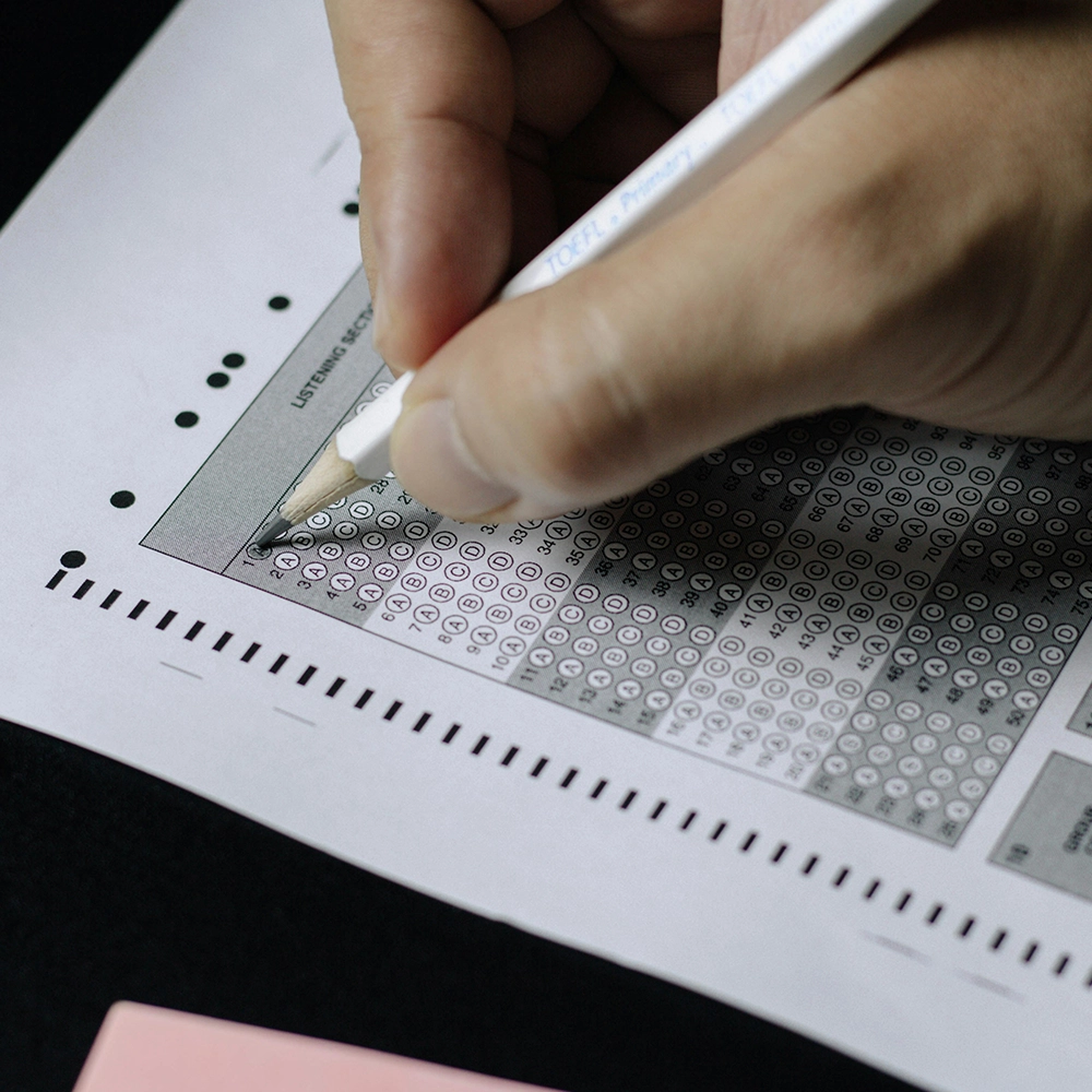 Op de foto zie je een hand die met een potlood antwoorden invult op een multiple-choice antwoordformulier, typisch voor gestandaardiseerde toetsen zoals de TOEFL. Het formulier bevat meerdere rijen met keuzemogelijkheden (A, B, C, D) die door de kandidaat kunnen worden ingevuld. De bubbels worden waarschijnlijk gescand door een machine. Links op het potlood staat "TOEFL Primary", wat aangeeft dat dit onderdeel is van een toets voor taalvaardigheid in het Engels.