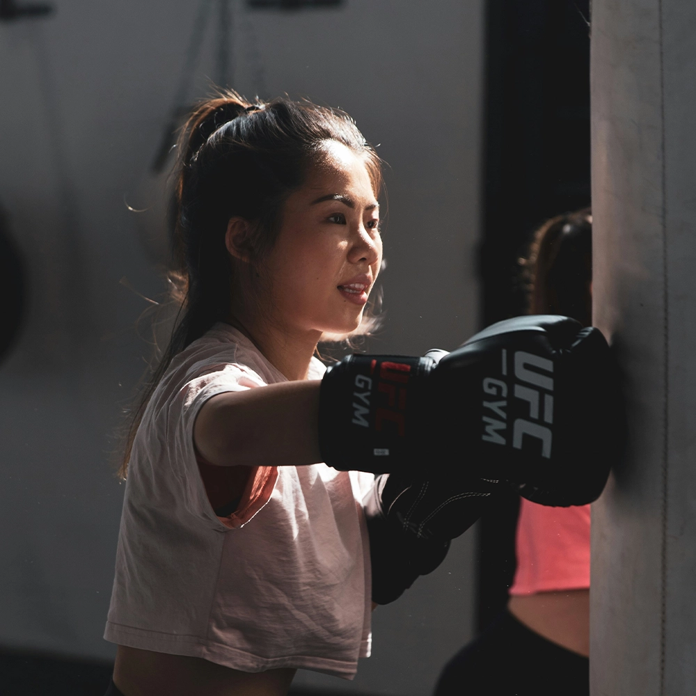 Op de foto zie je een vrouw die aan het boksen is in een sportschool. Ze draagt zwarte bokshandschoenen met het logo "UFC Gym" erop en slaat tegen een bokszak. Haar houding en gezichtsuitdrukking tonen focus en kracht. Het licht valt mooi op haar gezicht, wat het moment extra dynamisch maakt. Het lijkt een intensieve training te zijn in een professionele omgeving.