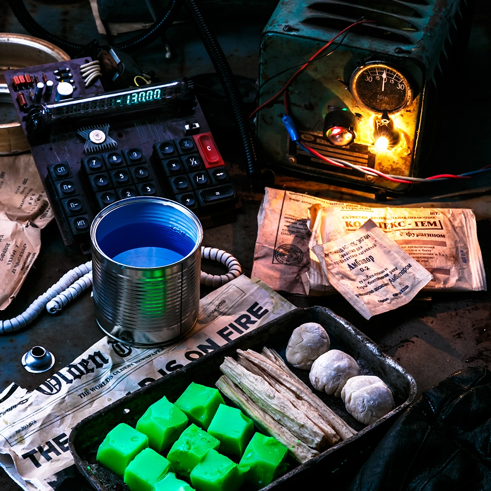 Een post-apocalyptisch tafereel toont een geïmproviseerde werkplek met militaire rantsoenen, een gloeiend oud apparaat, felgroene blokjes in een bak, een metalen blik met vloeistof en een krant met de kop "THE WORLD IS ON FIRE", wat samen een sfeer van chaos en overleven oproept.