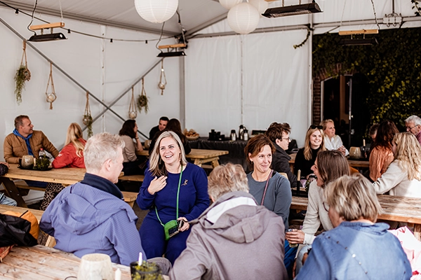 In een gezellige, met planten en lampionnen aangeklede tent zit een groep mensen ontspannen aan houten picknicktafels te kletsen, lachen en genieten van elkaars gezelschap.