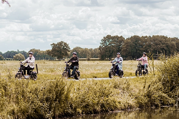 Vier mensen rijden ontspannen op elektrische fatbikes over een smal landweggetje door een groen, weids landschap met bossen op de achtergrond en een sloot op de voorgrond.