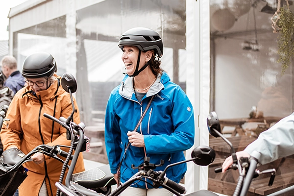 Twee vrouwen staan vrolijk naast hun elektrische fatbikes, klaar om te vertrekken, terwijl ze helmen dragen en gekleed zijn in kleurrijke regenjassen voor een avontuurlijke rit.