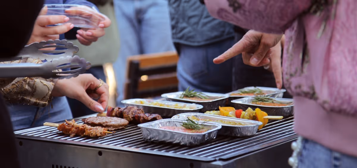 Mensen verzamelen zich rond een barbecue, terwijl ze samen genieten van gegrilde spiesen, hamburgers en gerechten in aluminium bakjes vol groenten en sausjes.