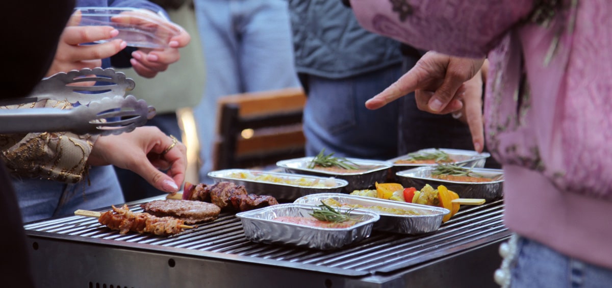 Mensen verzamelen zich rond een barbecue, terwijl ze samen genieten van gegrilde spiesen, hamburgers en gerechten in aluminium bakjes vol groenten en sausjes.